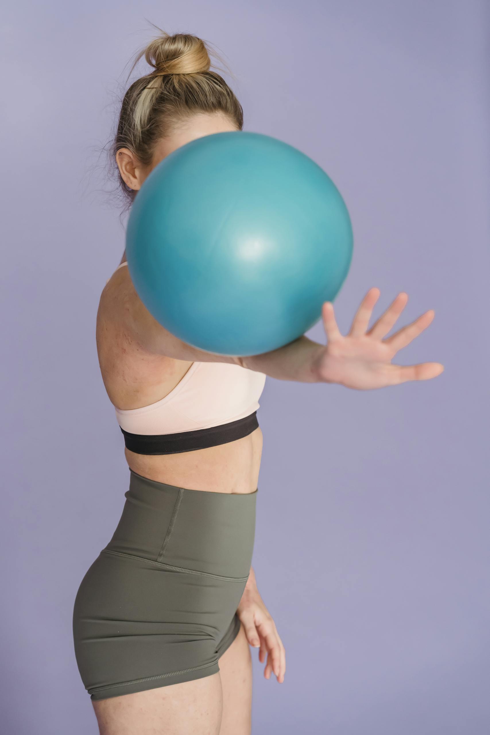 Faceless fit female in sportive top and shorts practicing with fitness ball while standing on purple background in modern studio