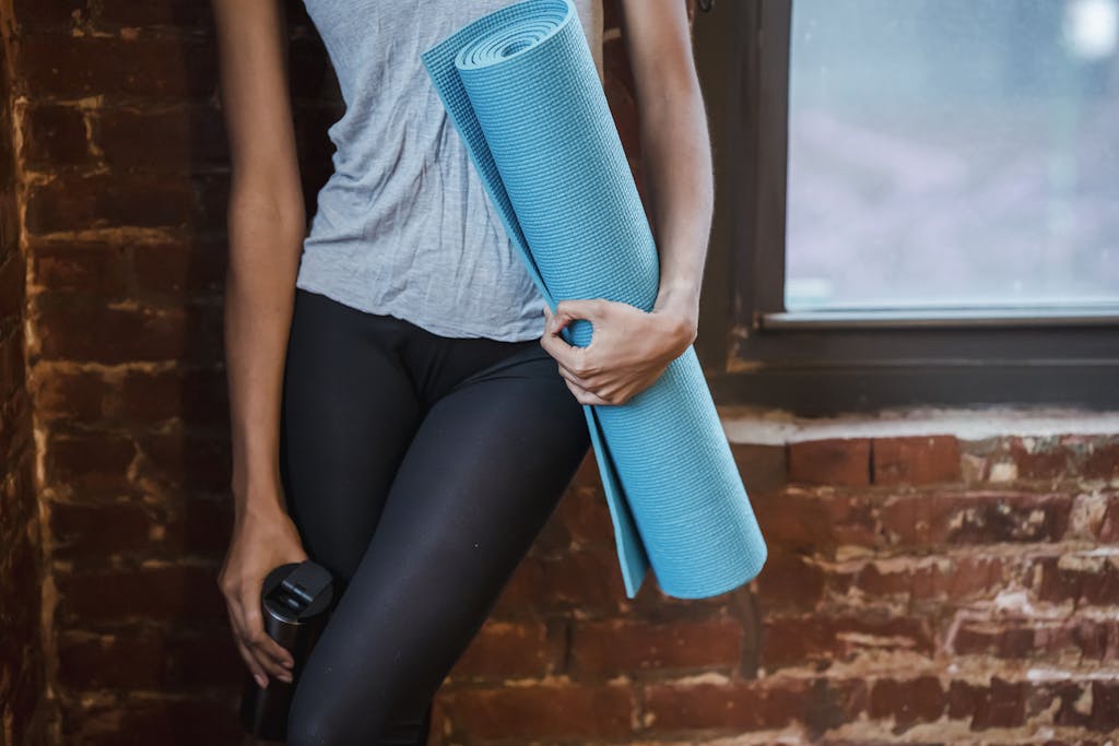 Unrecognizable sportive female in activewear standing near brick wall and window with rolled mat in hand during fitness training in gym