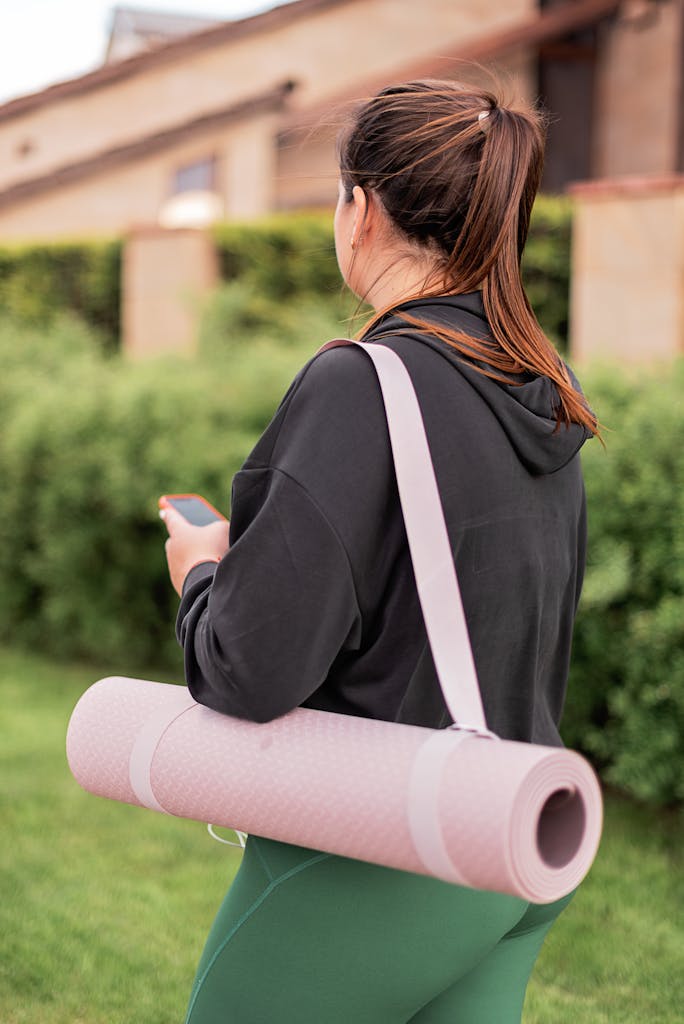 Woman carrying a pink yoga mat outdoors, wearing a black hoodie and green leggings.